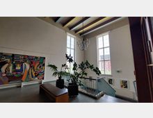 Innenraum eines Gebäudes mit hohen Decken, großen Fenstern und sichtbaren Holzbalken. An der Wand hängt ein farbenfrohes abstraktes Gemälde, daneben befinden sich Pflanzen, ein schlichtes Sitzmöbel sowie eine Treppe mit einem blauen Geländer; Kunstwerke sind entlang der Treppe aufgehängt.