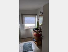 Kleines Badezimmer mit quadratischen beigen Fliesenboden, einem Holzwaschtisch mit schwarzem Waschbecken und Stauraum für Handtücher darunter. Ein Fenster mit halb heruntergelassenem Rollo und eine Glas-Duschkabine sind sichtbar.