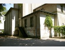 Außenansicht eines alten, verputzten Gebäudes mit sichtbaren Abnutzungsspuren und Graffiti an den Wänden, einer Metalltreppe zu einer erhöhten Tür sowie rankendem Grün an einer seitlichen Wand. Im Vordergrund ein asphaltierter Bereich, im Hintergrund Bäume, die Schatten spenden.