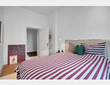 Schlafzimmer mit weiß gestrichenen Wänden und einem Bett mit rot-weiß gestreifter Bettwäsche. Daneben stehen ein Nachttisch mit Lampe und Büchern sowie ein gemaltes Bild, das an die Wand gelehnt ist. Im Hintergrund ist eine offene Zimmertür zu sehen.