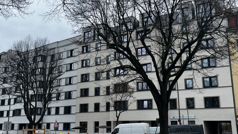Straßenszene mit einem langen, mehrstöckigen Wohn- oder Bürogebäude mit hellen Fassaden und vielen gleichmäßigen Fenstern, teilweise mit Dachgauben. Im Vordergrund stehen kahle Bäume sowie Baustellenabsperrungen und Transporter entlang der Straße.