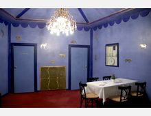 Raum mit blauen Wänden und goldenen Tiermotiven als Wanddekoration. Ein weiß gedeckter Tisch mit schwarzen Stühlen steht auf einem roten Teppich, ein Kronleuchter hängt an der Decke.