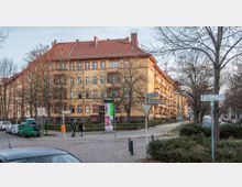 Eine Wohnhausanlage mit gelber Fassade und roten Fensterrahmen an einer Straßenecke, umgeben von kahlen Bäumen in einer städtischen Umgebung. Im Vordergrund befinden sich Schilder, geparkte Autos und ein Gehweg, auf dem Menschen zu sehen sind.