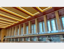 Weitläufige Halle eines Gebäudes mit hohen, rechteckigen Fenstern, die von beigefarbenen Säulen getrennt sind. Die Decke ist mit roten und gelb beleuchteten Paneelen gestaltet, und im unteren Bereich befinden sich Ticketschalter sowie ein Schild mit der Aufschrift „Der Tagesspiegel“.