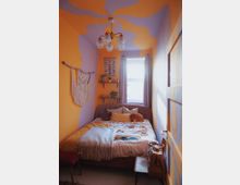 Schmales Schlafzimmer mit gelb-lila gestrichenen Wänden und Decken in einem organischen Muster. Im Raum stehen ein Bett mit gemusterten Kissen und einer Decke, ein Hängeregal mit Pflanzen und Dekoration, eine Wanddekoration aus Makramee sowie ein mittig hängender Lampenschirm mit Glasdetails; ein Fenster sorgt für Tageslicht.