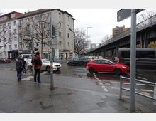 Straßenszene in einer urbanen Umgebung mit Mehrfamilienhäusern im Hintergrund, vorbeifahrenden Autos und einem erhöhten U-Bahn-Viadukt auf der rechten Seite. Personen stehen am Gehsteig, eine hält ein Fahrrad, während sie auf die Überquerung der Straße warten.