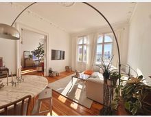 Helles Wohnzimmer mit hohen Decken, verzierten Stuckdetails und großen Rundbogenfenstern, die viel Tageslicht hereinlassen. Der Raum ist möbliert mit einem hellen Sofa, einem Glastisch und dekorativen Pflanzen, während ein angrenzender Raum durch eine breite Türöffnung sichtbar ist.