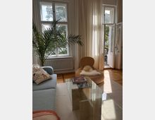 Wohnzimmer mit großen Fenstern und weißen Vorhängen, die natürliches Licht hereinlassen. Im Raum befinden sich ein hellblaues Sofa mit dekorativen Kissen, ein Glastisch, eine rattanartige Sitzgelegenheit mit Schaffell sowie eine Palme als Zimmerpflanze; im Hintergrund ist ein offener Zugang zu einem Balkon sichtbar.