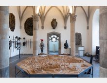 Museumsausstellung in einem historischen Raum mit gotischen Gewölbedecken und steinernen Säulen. Im Vordergrund ein detailliertes Holzmodell einer Stadt, im Hintergrund Ritterrüstungen, Schwerter und Wappenreliefs an den Wänden.