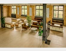 Großer Loft-Loungebereich in einem Industriebau mit freiliegenden Backsteinwänden, Stahlträgern und hohen Sprossenfenstern. Mehrere Sofagruppen mit niedrigen weißen Tischen stehen auf glattem Betonboden, ergänzt durch große Topfpflanzen und eine frei stehende Trennwand.