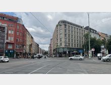 Straßenszene mit einer großen Kreuzung in einer Stadt. Zu sehen sind mehrstöckige Gebäude mit Restaurants und Geschäften im Erdgeschoss, Straßenbahnschienen, Fußgänger, Autos und eine U-Bahn-Station.