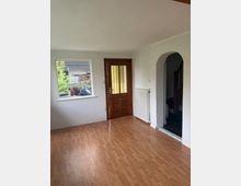 Leerer Innenraum mit Laminatboden, weiß gestrichenen Wänden und einer Holz-Eingangstür mit gelblich strukturierten Glasscheiben. Links befindet sich ein Fenster mit Blick nach draußen, rechts ein Rundbogen-Durchgang zu einem angrenzenden Bereich sowie ein Heizkörper an der Wand.