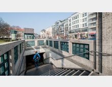 Eingang zu einer U-Bahn-Station in einer Stadtumgebung, gekennzeichnet durch ein Schild mit "U2 Pankow". Umgeben von Grünflächen, modernen Gebäuden mit Geschäften und parkenden Autos.