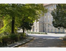 Historisches Gebäude mit heller Fassade und großen Fenstern, umgeben von einem Park mit Bäumen, Bänken und gepflasterten Wegen. Im Vordergrund sind schattige Sitzgelegenheiten unter Baumkronen zu sehen.