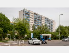 Mehrstöckiges Wohngebäude mit weißer Fassade und blauen Balkonen, umgeben von Bäumen und Grünflächen. Im Vordergrund befindet sich eine Straßenkreuzung mit parkenden Autos, einem Motorrollerfahrer und einem kleinen Kiosk.