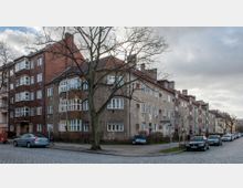 Straßenszene mit mehrstöckigen Wohngebäuden aus Backstein und Putzfassaden, einigen Bäumen im Vordergrund und geparkten Autos entlang der gepflasterten Straße. Der Himmel ist bewölkt, und es sind keine Personen im Fokus sichtbar.