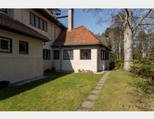 Haus im traditionellen Stil mit heller Putzfassade, roten Ziegeldächern und mehreren Fenstern mit Gittern. Im Vordergrund befindet sich ein Rasen mit einem gepflasterten Weg, umgeben von Bäumen und Sträuchern.