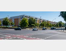 Straßenkreuzung in einer städtischen Umgebung mit mehreren Fahrspuren und Ampeln, im Hintergrund moderne mehrstöckige Wohngebäude in hellen Farben, flankiert von Bäumen und Grünflächen. Autos fahren und warten an der Kreuzung unter klarem Himmel.