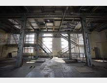 Industriehalle mit offenen Metalltreppen und Pfeilern, großen Fenstern, gelblichen Wandfliesen und sichtbaren Stahlträgern an der Decke. Der Raum ist leer, mit metallischem Boden und einem rustikalen, unbehandelten Erscheinungsbild.