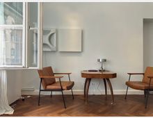 Minimalistisch eingerichteter Raum mit zwei braunen Ledersesseln, einem kleinen Holztisch mit geschwungenen Beinen und einer modernen Lampe. Im Hintergrund eine offene weiße Fensterfront, weiße Wände und abstrakte Wandkunst.