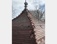 Dach eines Gebäudes mit roten Ziegeln, teilweise beschädigt oder fehlend, sowie ein kleiner Metallaufsatz an der Spitze. Im Hintergrund sind kahle Bäume und ein bewölkter Himmel zu sehen.