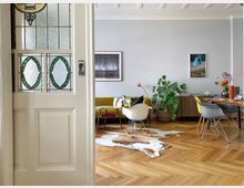 Wohnzimmer in einem Altbau mit Parkettboden und hohen Decken. Im Vordergrund eine Tür mit verziertem Glas, im Innenraum ein gelbes Sofa, ein Couchtisch, eine Zimmerpflanze in einem Korb, sowie ein Esstisch mit modernen Stühlen.