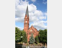 Backsteinkirche im neogotischen Stil mit hohem spitzen Turm und großen Fenstern, umgeben von Bäumen und einem niedrigen Zaun. Im Vordergrund eine Straße mit Fußgängern und Straßenlaternen, darüber ein blauer Himmel mit Wolken.