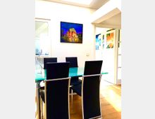 Heller Essbereich mit rechteckigem Glastisch und mehreren schwarzen Hochlehner-Stühlen auf Holzboden. An der weißen Wand hängt ein gerahmtes Bild, daneben führen Glastüren in angrenzende Räume mit farbigen Wandbildern.