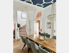 Esszimmer in einem modernen, hellen Wohnhaus mit einem großen Holztisch und grauen Polsterstühlen auf einem Fischgrät-Holzboden. Die Gestaltung umfasst eine blaue Decke mit weißen geometrischen Mustern, eine gläserne Kronleuchterlampe und eine geschwungene Holztreppe im Hintergrund.