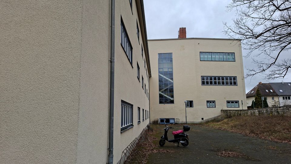 Hof zwischen zwei hell verputzten Mehrgeschossgebäuden mit vielen rechteckigen Fenstern und einem hohen, verglasten Treppenhaus. Der asphaltierte Platz ist von Laub bedeckt; am Rand stehen eine Laterne und eine niedrige Steinmauer mit angrenzenden Wohnhäusern im Hintergrund.