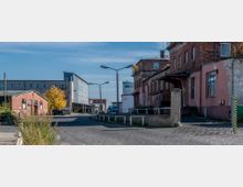 Straßenszene in einem Industriegebiet mit alten Backsteingebäuden und einem modernen grauen Gebäude im Hintergrund. Am Straßenrand stehen Laternen, und ein Baum mit gelbem Laub ist sichtbar.