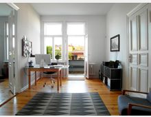 Helles Homeoffice mit Holzboden, einem Glastisch mit Computer und einem schwarzen Drehstuhl. Im Hintergrund sind große Fenster mit Blick auf einen Balkon und umliegende Gebäude, rechts ein Regal mit Pflanzen und links eine Pinnwand.