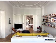 Bright, high-ceiling living room with ornate plaster crown molding, white walls, and a tiled floor, furnished with a yellow sofa, low white media console, and a TV. Long wall-mounted shelves filled with books line the right wall, and open double doors lead to a dining area with a table and wall gallery beyond.