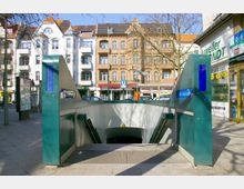 Eingang zur U-Bahn-Station „Bhf Schloßstraße“ mit seitlichen Rolltreppen, umgeben von Wohnhäusern mit historischer Fassade und Geschäften im Erdgeschoss in einer urbanen Umgebung.