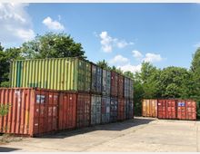 Mehrere gestapelte Frachtcontainer in verschiedenen Farben (Rot, Grün, Orange) stehen auf einem betonierten Lagerplatz, umgeben von Bäumen und blauem Himmel im Hintergrund.