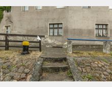 Eine beige, leicht beschädigte Hausfassade mit mehreren Fenstern, davor ein gepflasterter Gehweg mit steinernen Stufen. Im Vordergrund befinden sich ein Holzgeländer, ein gelb-schwarzer Pfosten und eine blaue Metallstange, die zwischen zwei Betonpfeilern befestigt ist.