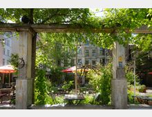 Gemütlicher Außenbereich eines Cafés oder Restaurants mit Holztischen, Metallstühlen, grünen Pflanzen und roten Sonnenschirmen, umgeben von alten Gebäuden und dekoriert mit Lichterketten. Im Vordergrund steht eine hölzerne Pergola, bewachsen mit grünem Laub.