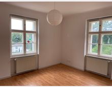 Leerer Raum mit hellem Laminatboden, weißen Wänden und zwei großen Fenstern mit Sprossen. Unter den Fenstern befinden sich Heizkörper, und an der Decke hängt eine runde Papierlampe. Draußen sind grüne Bäume und ein Nachbargebäude zu sehen.