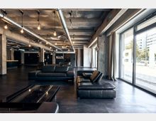Offener Innenraum mit industriellem Design, bestehend aus freiliegenden Betondecken, Rohrleitungen und großen Fenstern. Die Einrichtung umfasst schwarze Ledersofas, Glastische und einen dunklen Bodenbelag, mit Blick ins Freie auf moderne Gebäude.