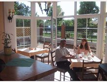 Ein heller Wintergarten mit großen bodentiefen Fenstern und Blick auf einen Garten mit Bäumen und Zaun. Zwei Personen sitzen an einem Tisch mit einer weißen Tischdecke und genießen eine Mahlzeit, während Sonnenlicht den Raum erhellt.