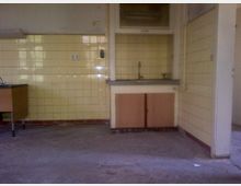 Ein leerer Raum mit einer Küchenzeile, bestehend aus einer Spüle mit zwei Holzschranktüren darunter. Die Wände sind mit gelben Fliesen verkleidet, der Boden ist unbedeckt und zeigt Beton. Links im Bild steht ein Tisch mit einer dunklen Arbeitsplatte und Metallbeinen.