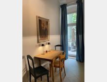 Kleiner Raum mit einem Holztisch, zwei Stühlen und zwei kleinen Tischlampen. An der Wand hängt ein gerahmtes Gemälde, und im Hintergrund ist eine Balkontür mit blauen Vorhängen zu sehen, die auf einen Außenbereich führt.