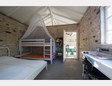 Ein Schlafzimmer mit freiliegenden Ziegelwänden, einem Doppelbett und einem Etagenbett mit Moskitonetz. Im Hintergrund ist eine geöffnete Tür, die in einen hellen Raum mit weiß gestrichenen Wänden und grünen Stühlen führt.