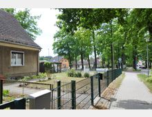 Wohngebiet mit Einfamilienhäusern neben einer mit Bäumen gesäumten Straße. Ein gefliestes Gehwegstück und ein Zaun trennen die Grundstücke von der Straße; im Vordergrund ein Garten vor einem Wohnhaus.
