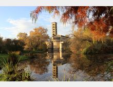 Historische Kirche mit Campanile im italienischen Stil, gelegen an einem ruhigen Teich, umgeben von herbstlichem Laub und Grasflächen. Die Reflektion des Gebäudes und der Bäume spiegelt sich klar im Wasser wider.