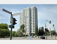 Hohes Wohngebäude mit klaren, modernen Linien an einer Kreuzung mit Verkehrsampeln und Straßenschildern, umgeben von Bäumen und vereinzeltem Verkehr; Straßenschilder weisen auf die "Breite Straße" und "Schopenhauerstraße" hin.