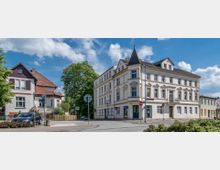 Straßenszene mit einem historischen Gebäude an einer Ecke, das drei Stockwerke hoch ist und eine helle Fassade mit dekorativen Elementen und einem kleinen Balkon besitzt. Links daneben steht ein modernes Wohnhaus mit Garten, und im Hintergrund sind Gehwege, Bäume und ein klarer Himmel zu sehen.