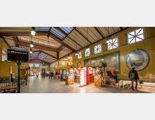 Innenraum eines historischen U-Bahnhofs mit gelb gekachelten Wänden, großen Fensterflächen und einem gewölbten Dach mit Stahlträgern. Es gibt Stände mit Verkaufsbuden, eine Anzeigetafel für Gleis III und Hinweisschilder für die Linie U1 Richtung Warschauer Straße.