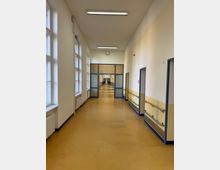 Langer, heller Flur in einem Gebäude mit gelblich-braunem Boden, gelb-weiß gestrichenen Wänden und blauer Türrahmung. Links befinden sich große Fenster mit Heizkörpern darunter, rechts mehrere Zimmertüren mit Handläufen an den Wänden.