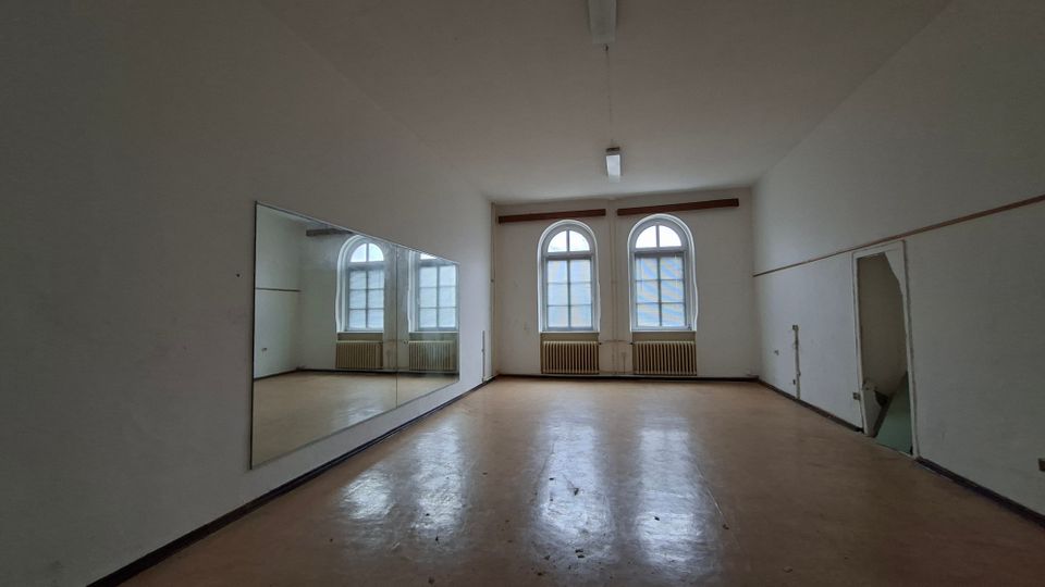 Leerer, rechteckiger Innenraum mit hellem Boden, weißen Wänden und zwei hohen Rundbogenfenstern an der Stirnseite, darunter Heizkörper. An der linken Wand hängt ein großer Spiegel, an der Decke sind Leuchtstofflampen, rechts befindet sich eine offene Türöffnung.